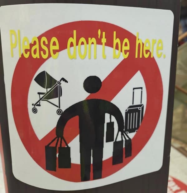 aggressive translation fail on a warning sticker. Featuring a crossed-out pictogram of a person laden with strollers, suitcases, and shopping bags, the text above simply commands: "Please don't be here."