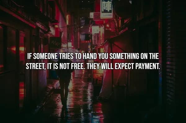 A travel hack regarding street safety over a rainy, neon-lit alleyway in a bustling city. The text warns: IF SOMEONE TRIES TO HAND YOU SOMETHING ON THE STREET, IT IS NOT FREE. THEY WILL EXPECT PAYMENT. This is a common warning for tourists to avoid high-pressure street peddler scams.