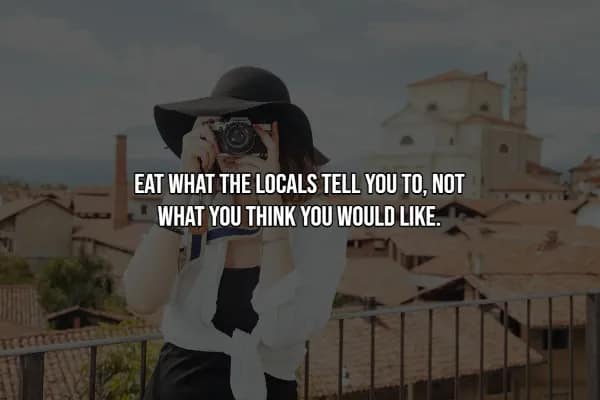 A travel hack advising tourists to trust local food recommendations over their own preferences. A woman in a wide-brimmed black hat holds a camera up to her face, with a terracotta-roofed Mediterranean town in the background.