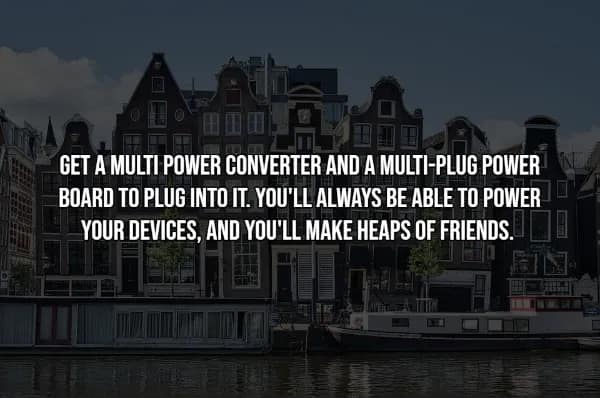 A travel hack for international charging over a picturesque view of traditional Dutch canal houses in Amsterdam. The text reads: GET A MULTI POWER CONVERTER AND A MULTI-PLUG POWER BOARD TO PLUG INTO IT. YOU'LL ALWAYS BE ABLE TO POWER YOUR DEVICES, AND YOU'LL MAKE HEAPS OF FRIENDS. This strategy allows for multiple devices to be charged from a single foreign outlet.