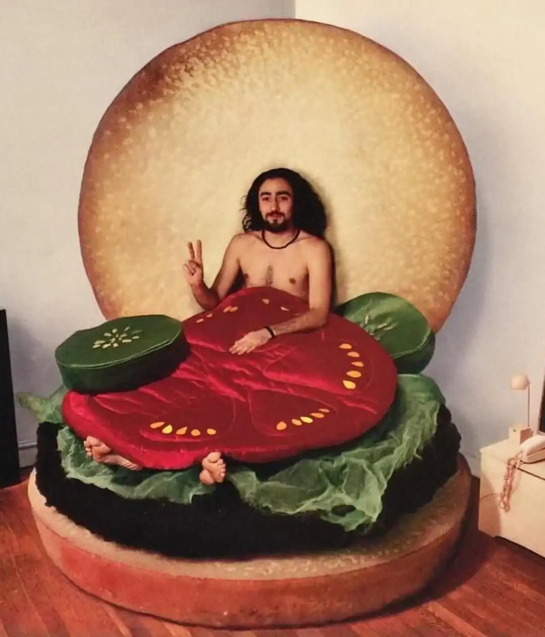 Shirtless man sitting inside giant hamburger bed with bun headboard lettuce tomato and pickle layers
