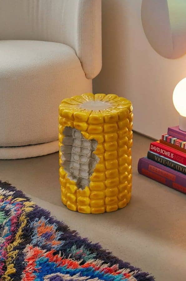 Ceramic yellow corn on the cob stool with bite missing sitting next to books and lamp