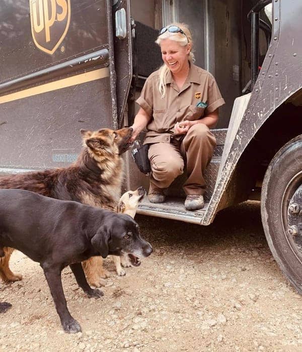 Female UPS driver sits on truck step petting three dogs on rural dirt road