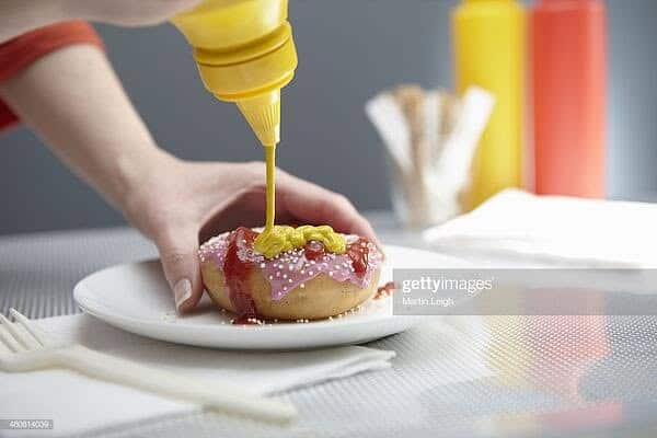 Strange weird stock photo of hand squeezing yellow mustard onto pink frosted sprinkled donut