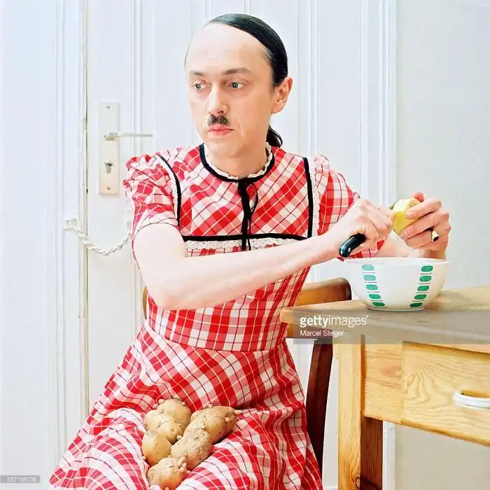 Bizarre weird stock photo of mustached man wearing red plaid dress peeling potatoes in kitchen