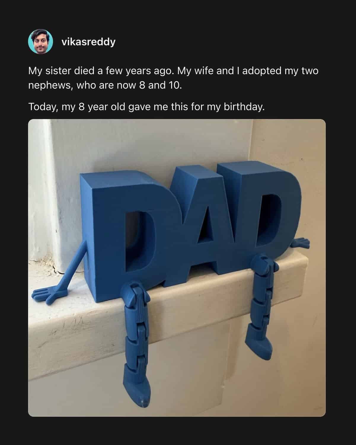 A heart-wrenching wholesome meme post features a man who adopted his nephews after his sister passed away. He shares a photo of a blue 3D-printed sign that spells out DAD with little legs dangling off the shelf, a birthday gift from his eight-year-old.