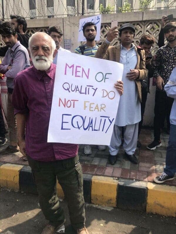 A powerful wholesome meme photograph of an elderly man with a long white beard and purple shirt standing at a protest. He holds a hand-lettered sign that reads: "MEN OF QUALITY DO NOT FEAR EQUALITY," showcasing an ally in the wild.