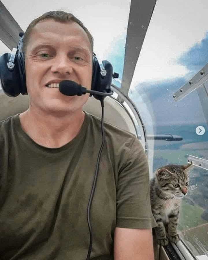 Tabby cat sitting in small aircraft cockpit beside smiling pilot during flight looking unimpressed