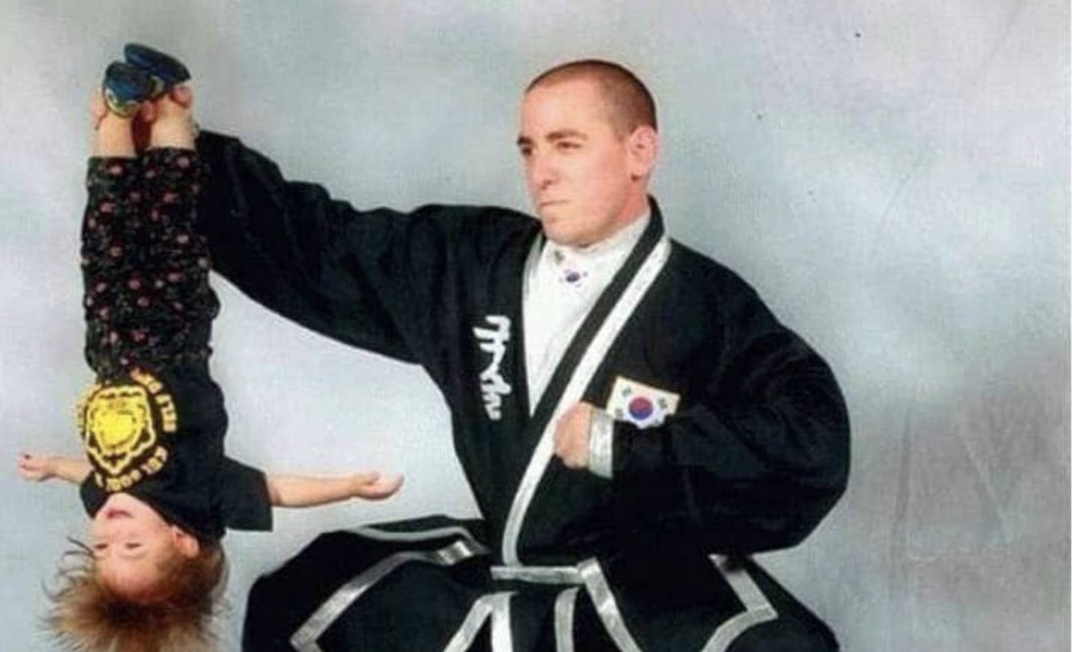 A martial arts instructor in a black uniform holds a toddler upside down by the feet.