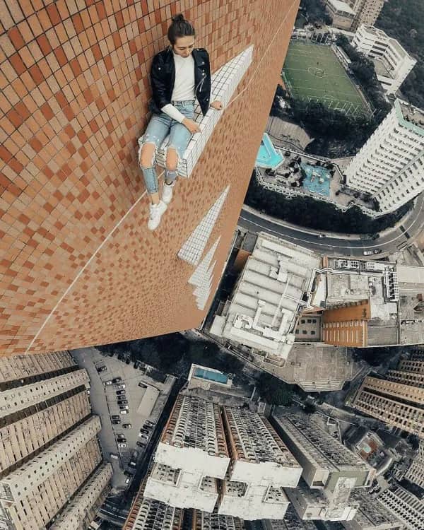 Woman with blonde hair sitting calmly on a narrow building ledge, overlooking a city.