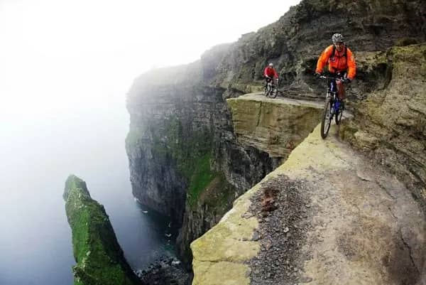 Two cyclists cautiously riding mountain bikes along a narrow, sheer coastal cliff edge.