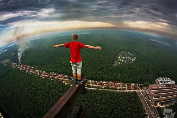 A person standing on a narrow metal beam, high above a dense forest.