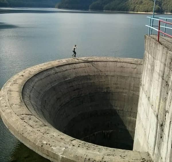 Long-distance view of a person unicycling precariously on the edge of a high concrete dam spillway.