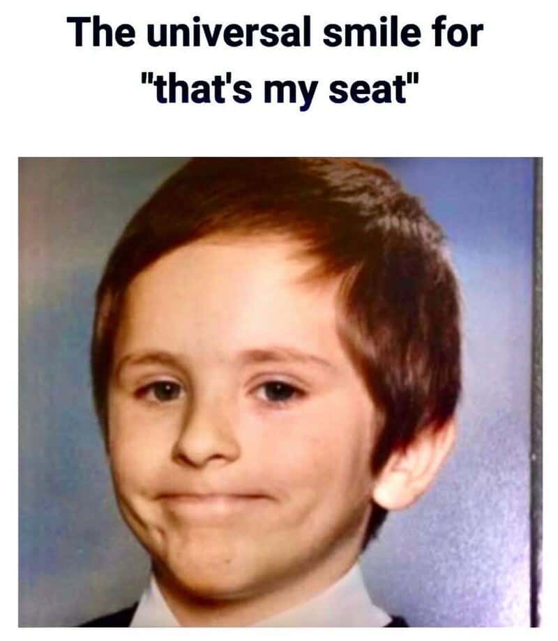 Close-up of a child’s awkward, tight-lipped smile captioned as the universal "that’s my seat" face.