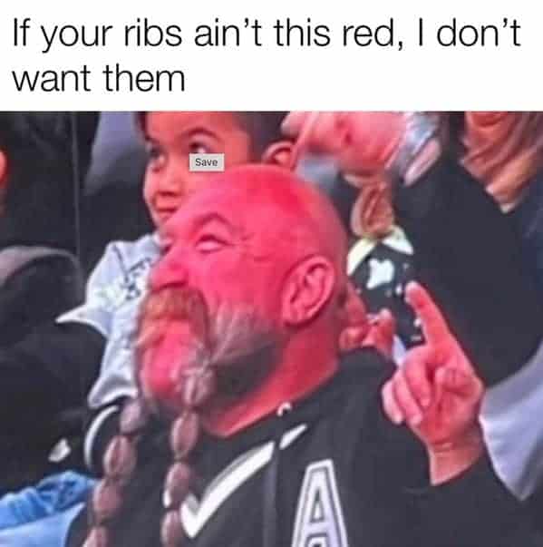 A candid stadium fan photo operates as a funny bbq meme, highlighting a bald man with a braided beard whose skin is cast in an intensely bright red neon light, captioned that if your smoked ribs aren't that color, they aren't wanted.