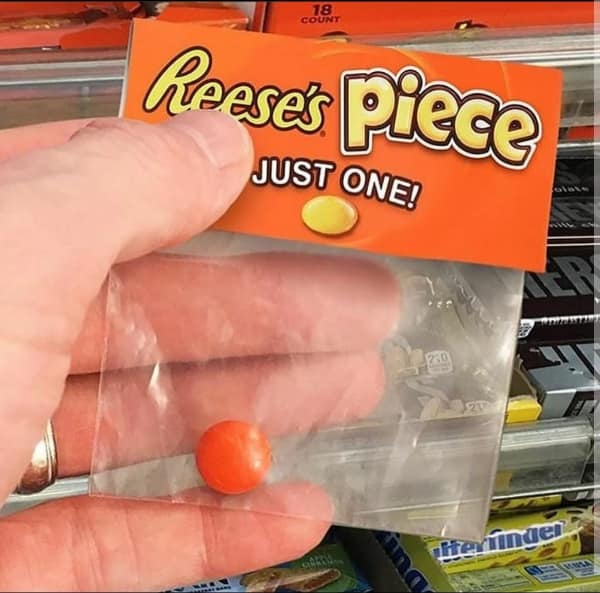 A retail product package features an orange cardboard header labeled "Reese's Piece" with the tag "JUST ONE!" sealing a miniature plastic ziplock bag containing a single orange candy in a funny blursed image.