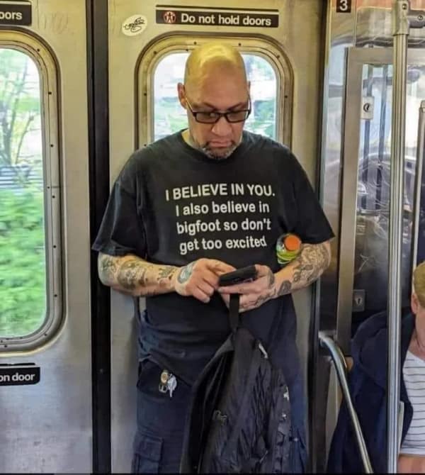 A heavily tattooed commuter standing on a train car wears a black t-shirt displaying a backhanded inspirational message about believing in Bigfoot, making for a popular blursed image.