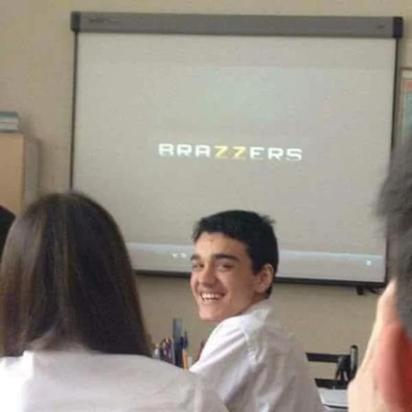 A high school classroom scene captures a student grinning back at a camera while a prominent adult website logo is accidentally projected onto the large whiteboard presentation screen in a funny blursed image.