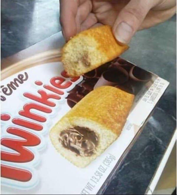 A close-up snapshot of a chocolate-filled snack cake broken open showcases an uneven manufacturing defect where the dark core filling is entirely missing from one end, featured as a textbook blursed image.