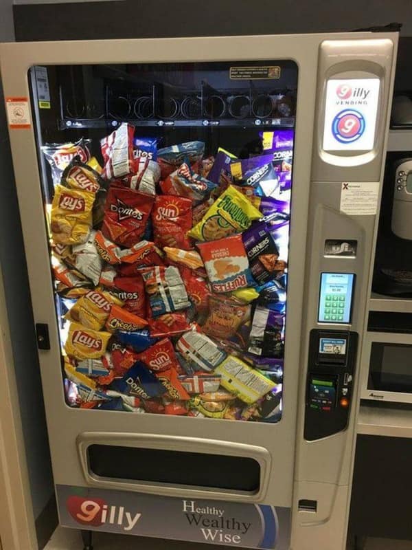A commercial glass-front vending machine packed solid to the top with un-dispensed bags of chips, Doritos, and Cheetos creates a chaotic blursed image of mechanical logistics failure.