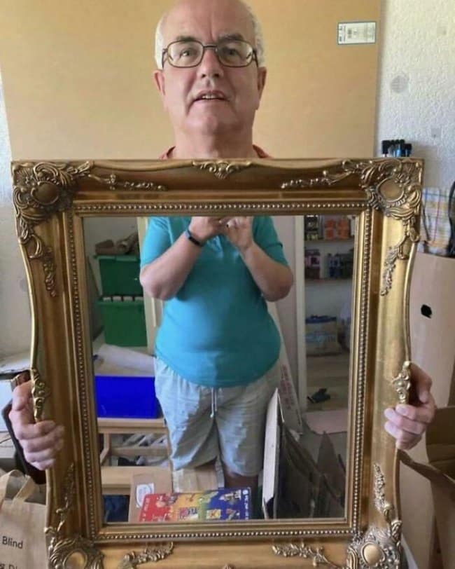 A photo of an older man holding up a large, ornate gold-framed mirror for sale. Due to the angle of the mirror and his height, his actual head is visible above the frame, while his reflected torso appears inside the frame, creating a disorienting "severed head" visual.