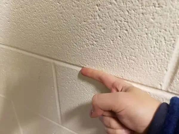 A close-up of a hand carefully sliding a finger along the deep, recessed grout lines of a painted cinder block school wall captures a highly specific tactile childhood meme.