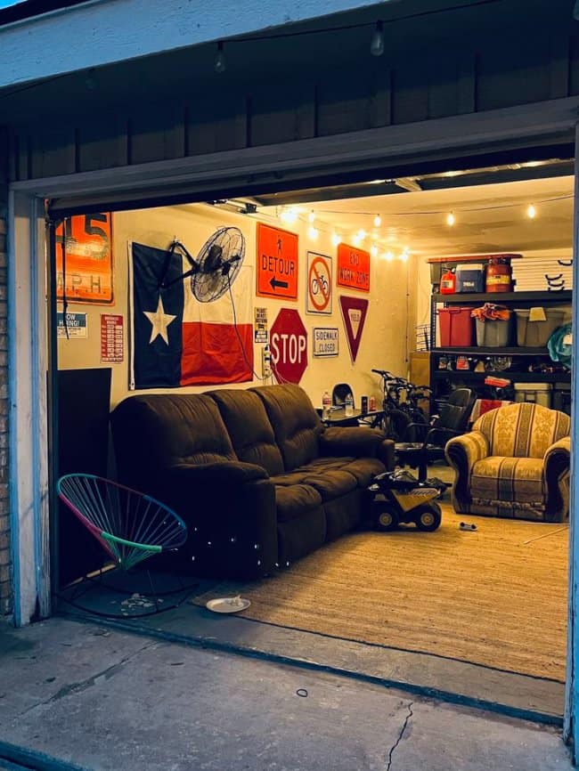A quintessential chill spot for dudes inside a suburban garage, featuring mismatched plush couches, a Texas state flag, stolen road signs for wall art, and warm string lights.