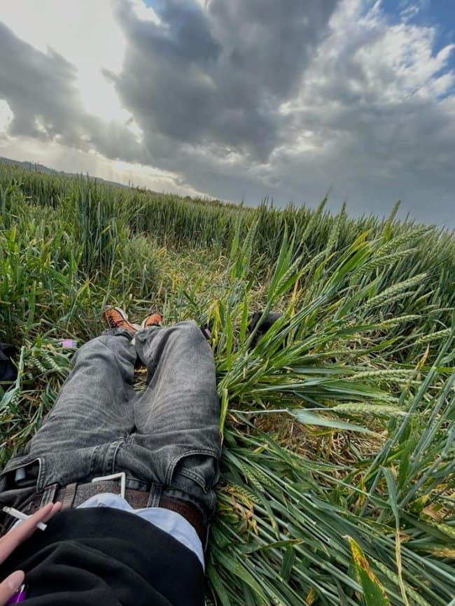 A pastoral chill spot for dudes from a POV perspective, lying down in a field of tall green wheat under a dramatic, cloudy sky while holding a cigarette.