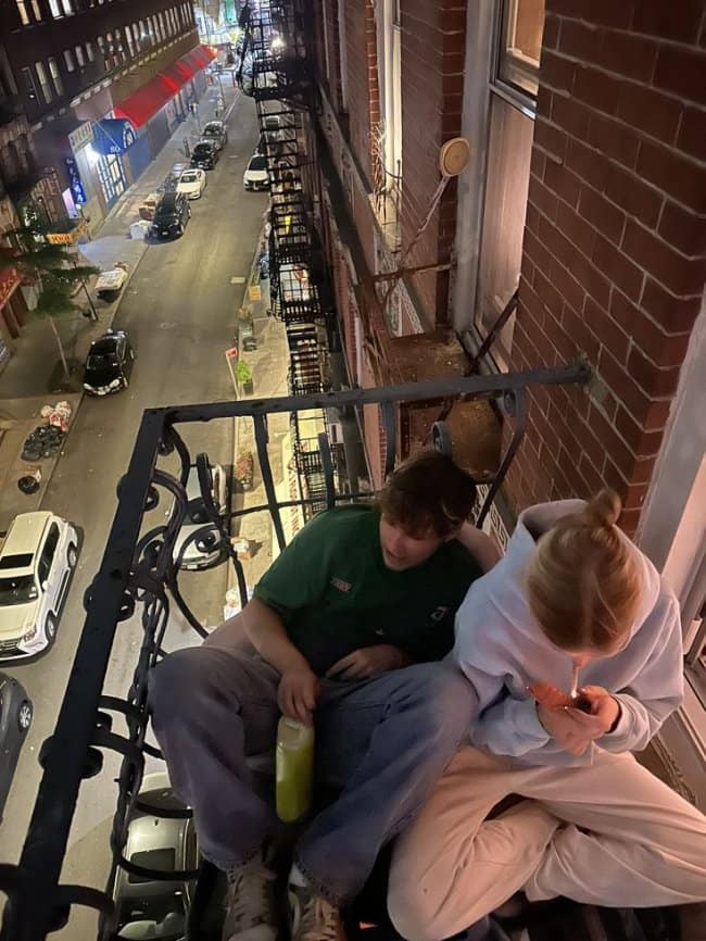 An atmospheric urban chill spot for dudes featuring two people sitting on a narrow iron fire escape at night, overlooking a glowing city street with red awnings and parked cars.