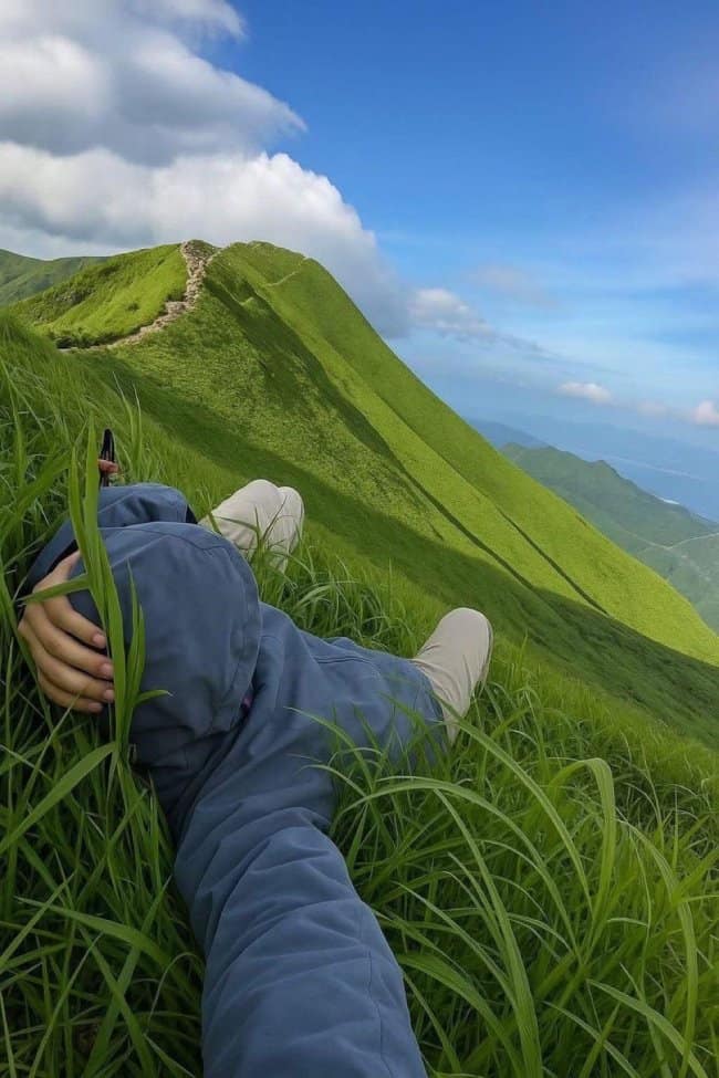 A breathtaking natural chill spot for dudes where the viewer is lying on a steep, vibrantly green grassy mountain ridge that overlooks a vast valley and distant peaks.