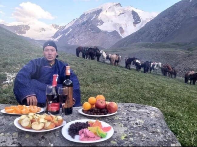 A legendary chill spot for dudes featuring a man in traditional Mongolian robes enjoying a sophisticated spread of red wine and fresh fruit on a rock, with horses and snow-capped peaks in the background.