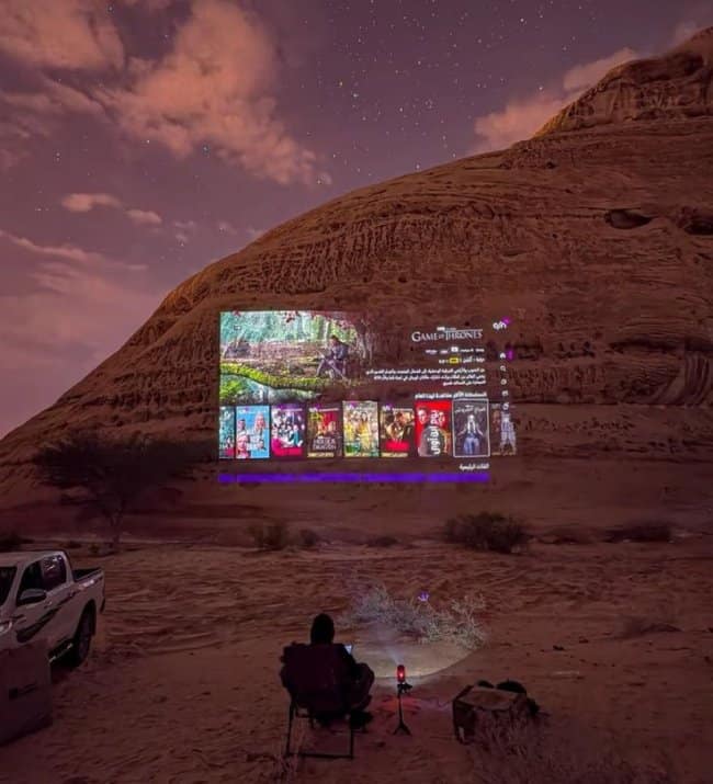 An elite outdoor chill spot for dudes showing a high-quality movie projector beaming a streaming menu onto the side of a massive desert rock formation under a starry night sky.