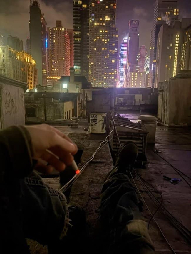 A gritty city chill spot for dudes from a POV of sitting on a rooftop edge at night, holding a lit cigarette with the glowing skyscrapers of a major metropolis in the background.