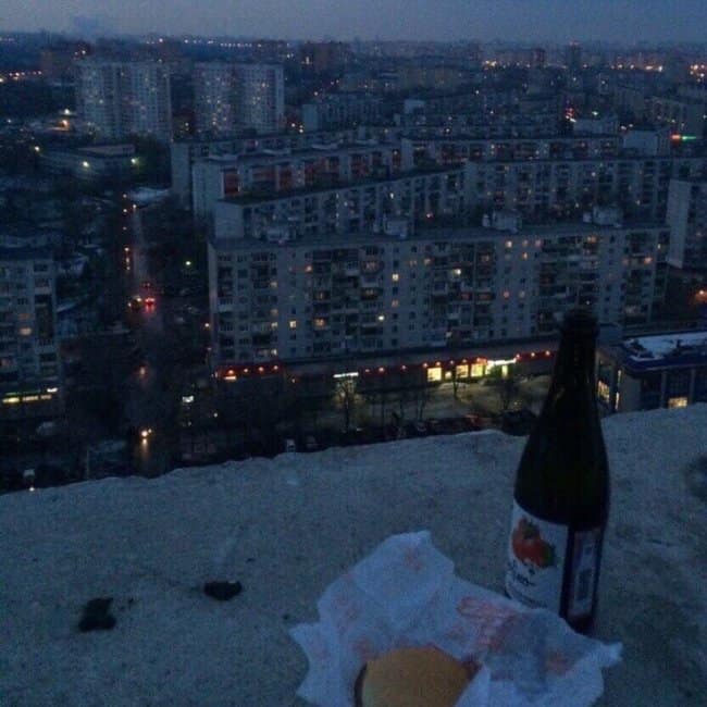 A somber but cozy chill spot for dudes on a cold concrete rooftop, featuring a lone bottle of beer and a burger overlooking a sprawling Eastern European apartment complex at twilight.