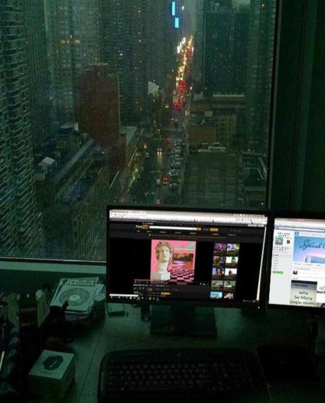 A moody chill spot for dudes featuring a dual-monitor desktop setup by a window overlooking a rainy city street at night, with the glowing tail lights reflecting off the wet pavement.