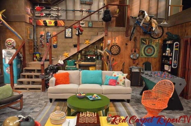 A maximalist chill spot for dudes in a wood-paneled basement, overflowing with skateboards, skis, a foosball table, a bicycle mounted to the wall, and bright orange accent chairs.