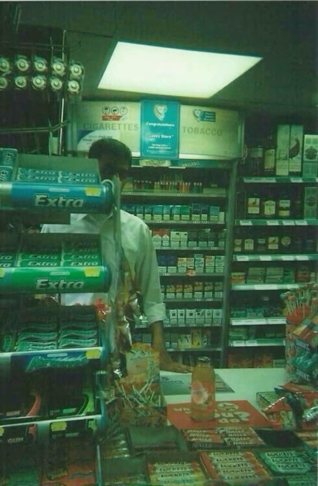 A nostalgic, liminal chill spot for dudes inside a dimly lit local convenience store at night, packed with rows of gum, tobacco, and candy, creating a quiet urban sanctuary.