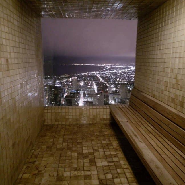 A high-end, modern chill spot for dudes inside a tiled sauna-style balcony overlooking a sprawling city skyline at night through a massive open window.