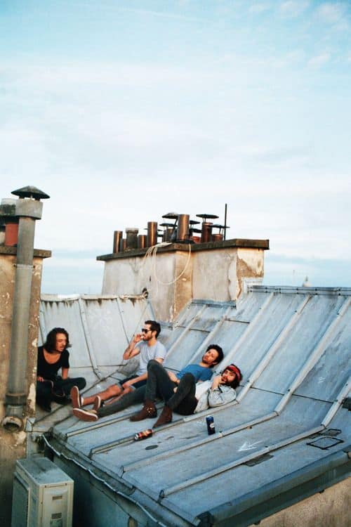 An improvised urban chill spot for dudes where four friends are lounging on a slanted metal rooftop between brick chimneys, enjoying drinks under an overcast sky.