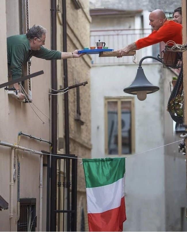 A legendary neighborly chill spot for dudes in a narrow European alleyway, where two men are passing a tray of espresso between windows using a long wooden board.