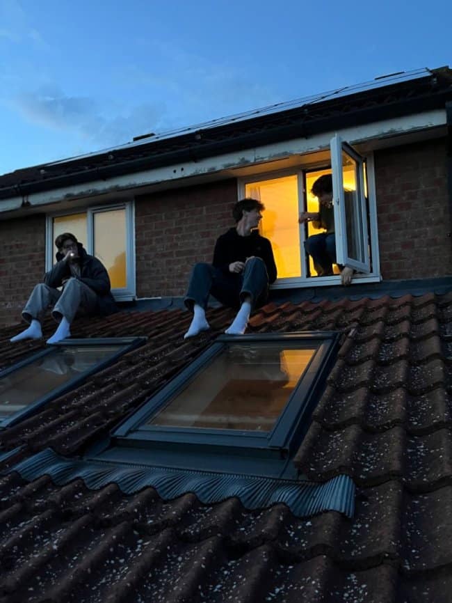A nostalgic blue-hour chill spot for dudes showing three friends sitting on the shingles of a house roof outside an open attic window.