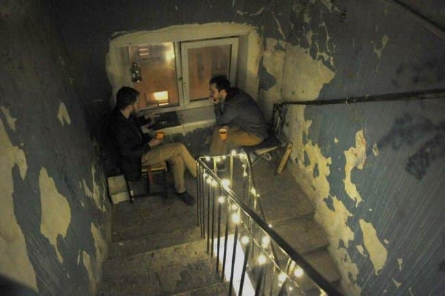 A liminal and cozy chill spot for dudes set up in a dilapidated stairwell, decorated with fairy lights and featuring two men having a deep conversation over coffee.