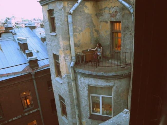 A solitary and scenic chill spot for dudes on a tiny, rounded concrete balcony of a crumbling vintage apartment building overlooking a European city.