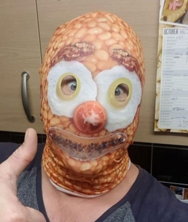 A surreal culinary photo serves as a bizarre classic meme featuring a person giving a thumbs-up while wearing a balaclava ski mask printed with a full English breakfast pattern made of baked beans, fried egg eyes, and a tomato nose.