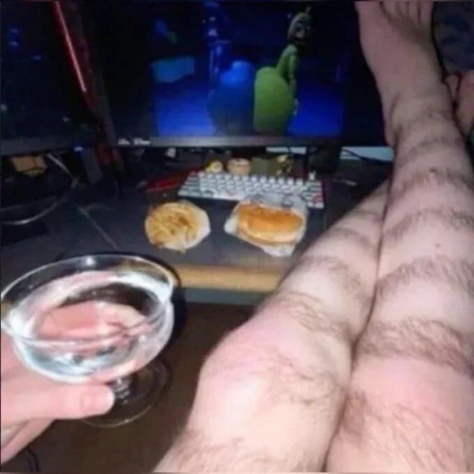 A cursed classic meme image showing a person's incredibly hairy legs resting on a desk with a gaming monitor in the background, a burger and fries on the desk, and a hand holding a glass of water, evoking a chaotic "gamer lair" aesthetic.
