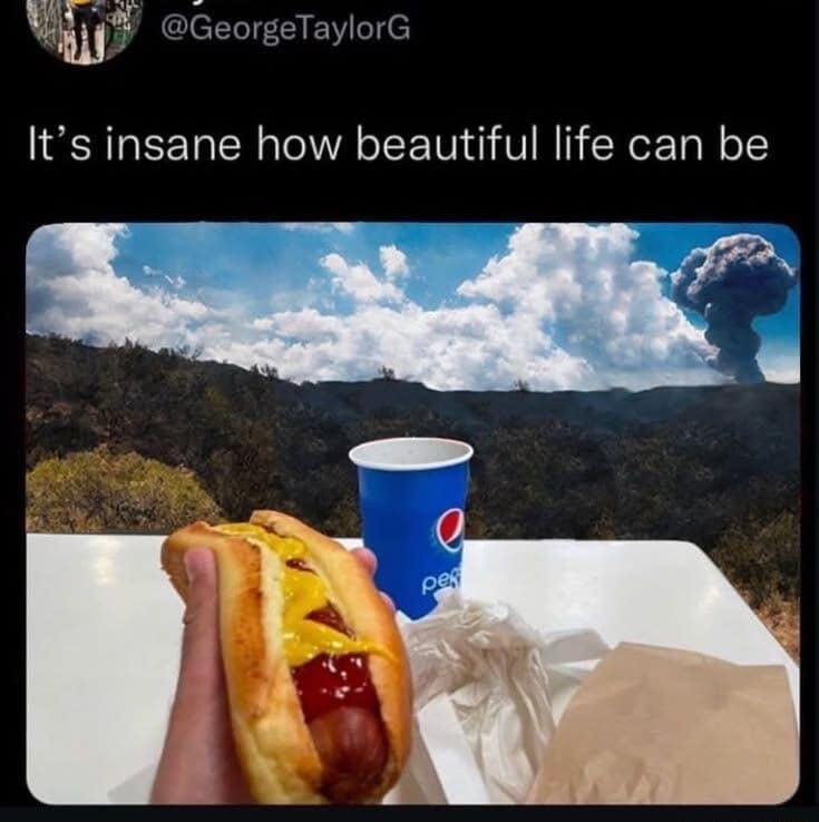 classic meme tweet showing a first-person perspective of someone holding a hot dog with ketchup and mustard next to a Pepsi cup on a white table, while a massive, dark volcanic mushroom cloud erupts violently on the horizon under a bright sky.