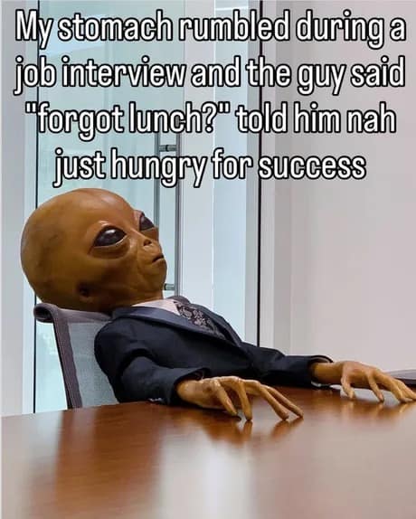 A classic meme featuring a grey alien in a sharp business suit sitting at a boardroom table, with a caption about playing off a stomach rumble in a job interview as being "hungry for success."