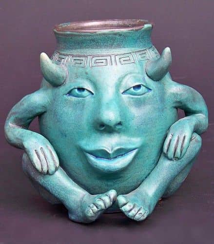 A teal ceramic jar featuring a human face with horns, sitting in a cross-legged yoga pose.