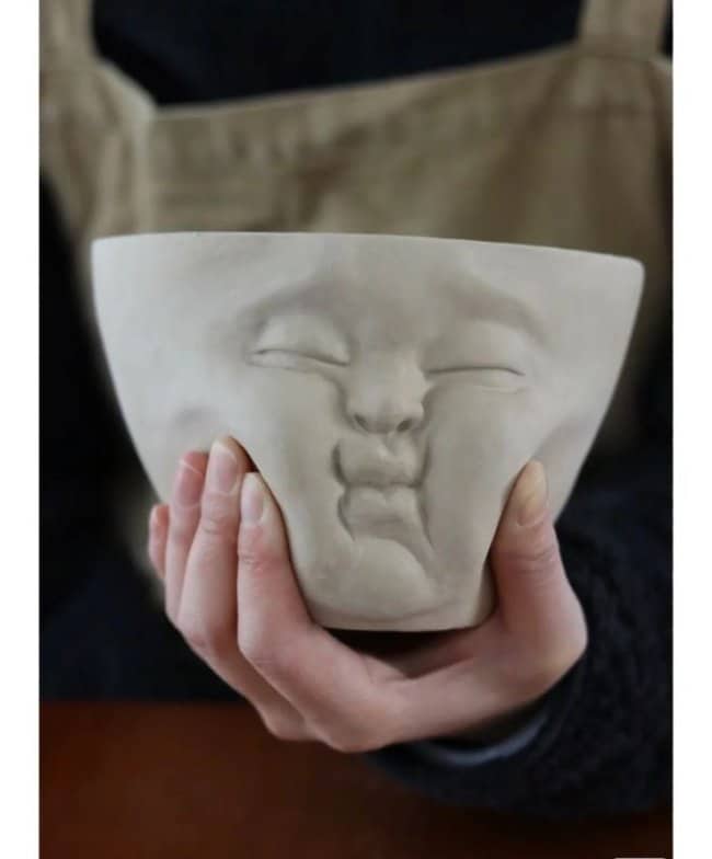 A person squeezing a malleable-looking ceramic bowl that features a squished, puckered human face.