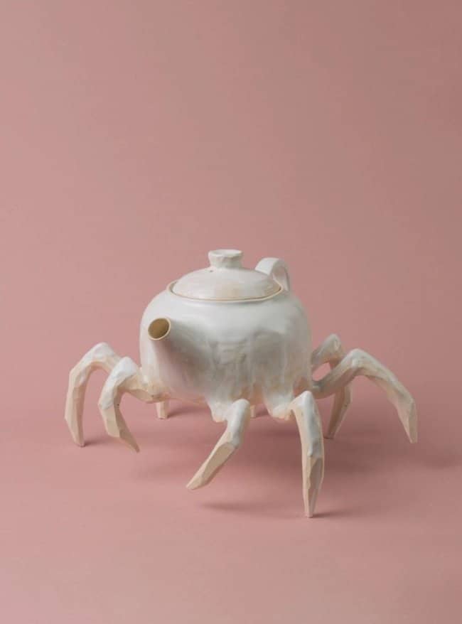 A white glazed teapot designed with eight sharp spider-like legs scuttling on a pink background.